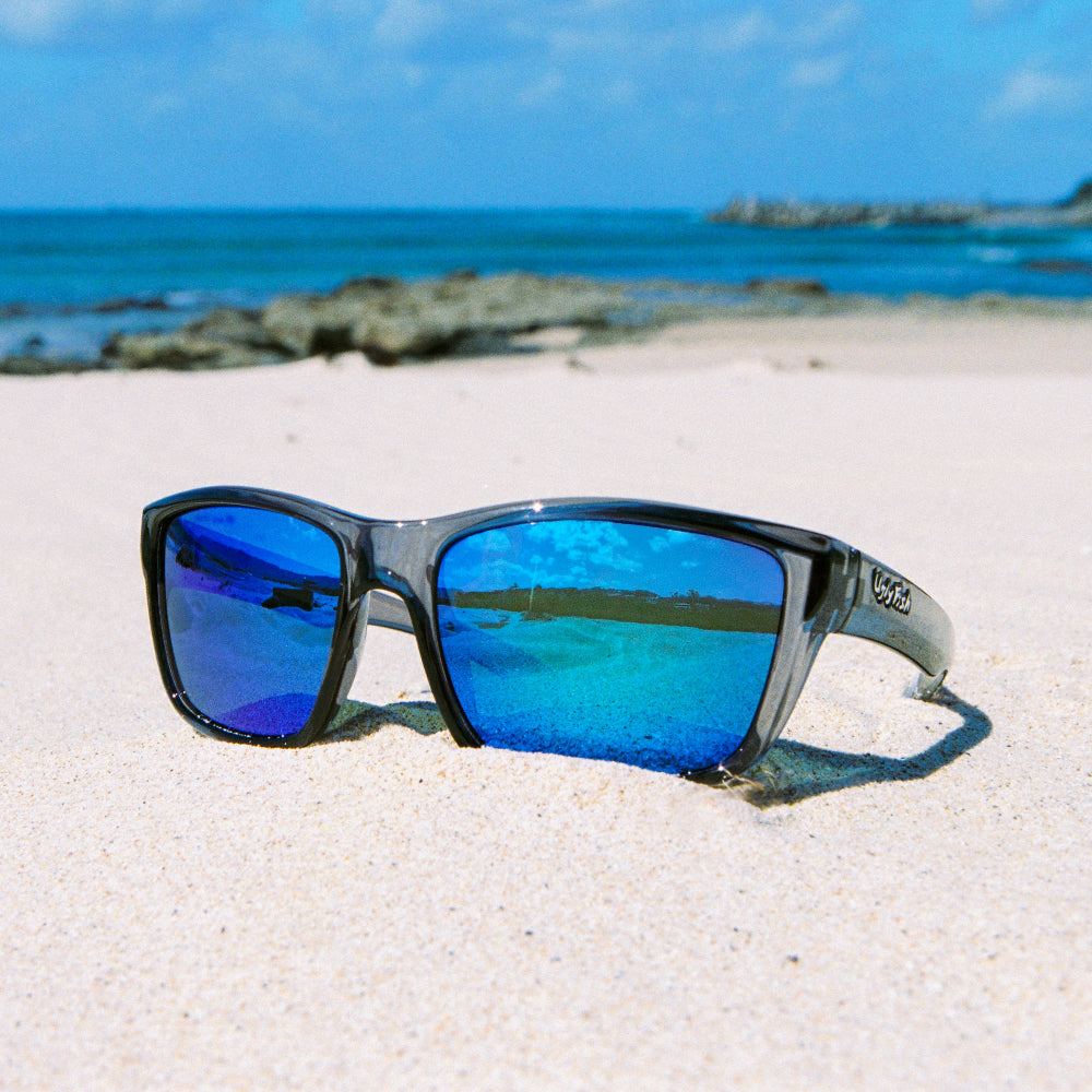Ugly Fish polarised sunglasses with blue lenses on a sandy beach with ocean in the background