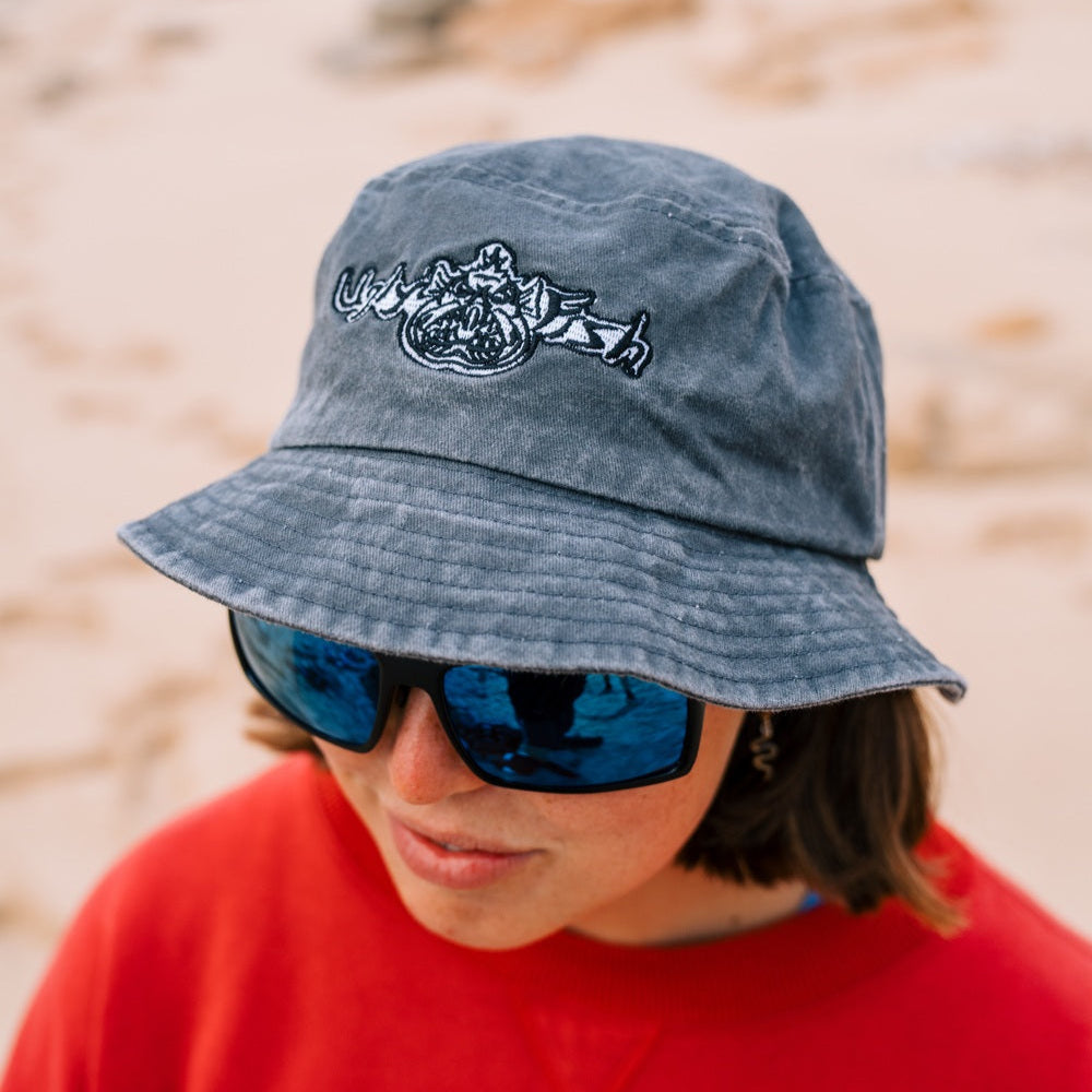 Person wearing a grey Ugly Fish bucket hat with embroidered logo on a beach.