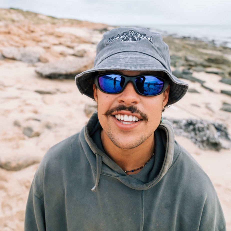Person wearing a grey Ugly Fish bucket hat and sunglasses with a beach background