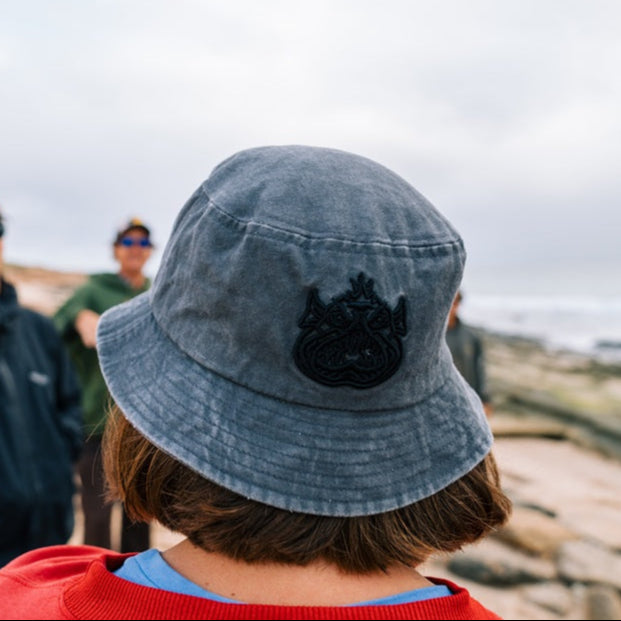 Person wearing a grey Ugly Fish bucket hat with a black logo, standing on a rocky landscape.