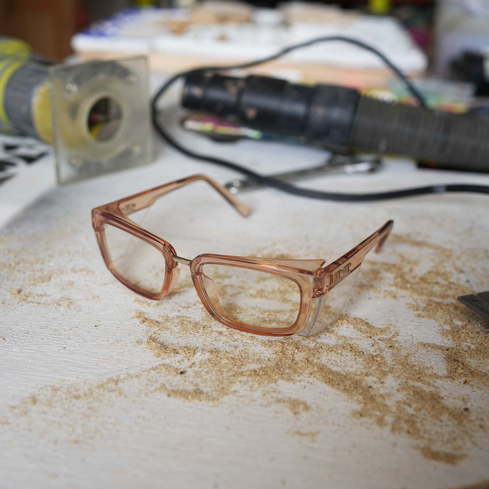 Champagne colored Ugly Fish safety eyeglasses on a surface with dust and tools in the background.