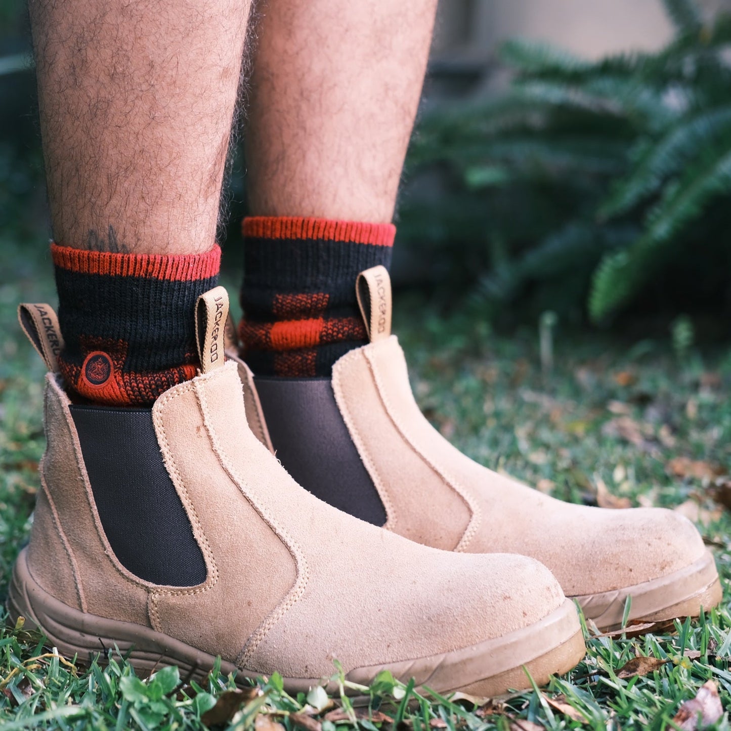 Person wearing beige work boots and black and red Ugly Fish socks on grass.