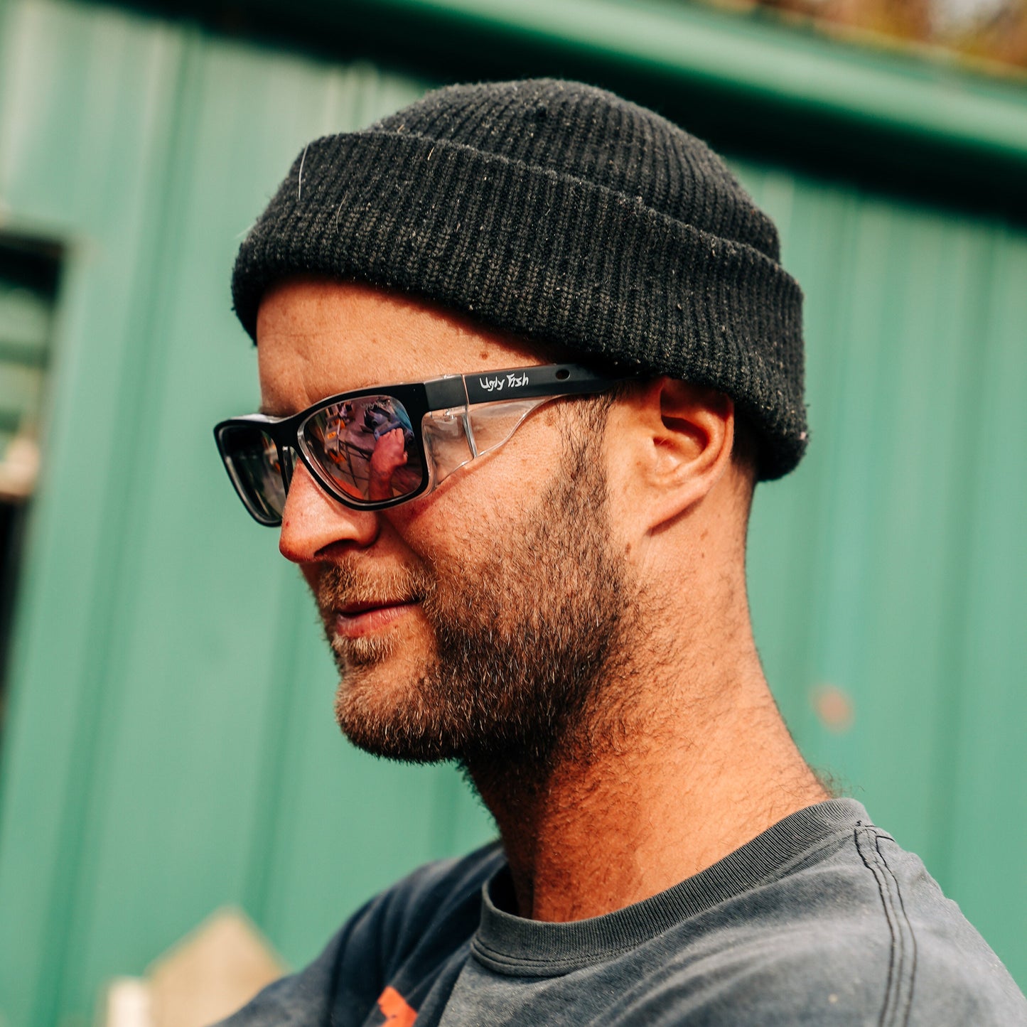 Man wearing a black beanie and sunglasses with a green metal fence in the background