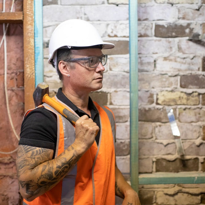 Person wearing a hard hat and Ugly Fish safety glasses holding a hammer in a construction setting.