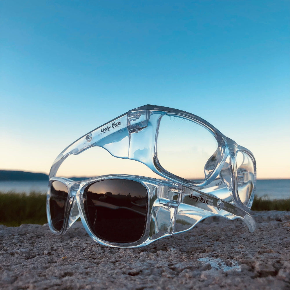 Clear Ugly Fish Safety sunglasses with photochromic lenses on a sandy surface with a blue sky and water in the background