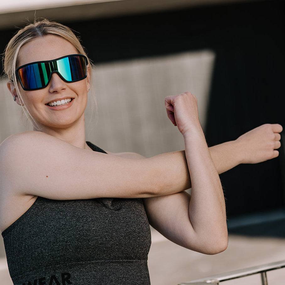 A woman in a dark gray sports bra and mirrored ugly fish sunglasses stretches her arm, standing outdoors in front of a modern concrete wall.