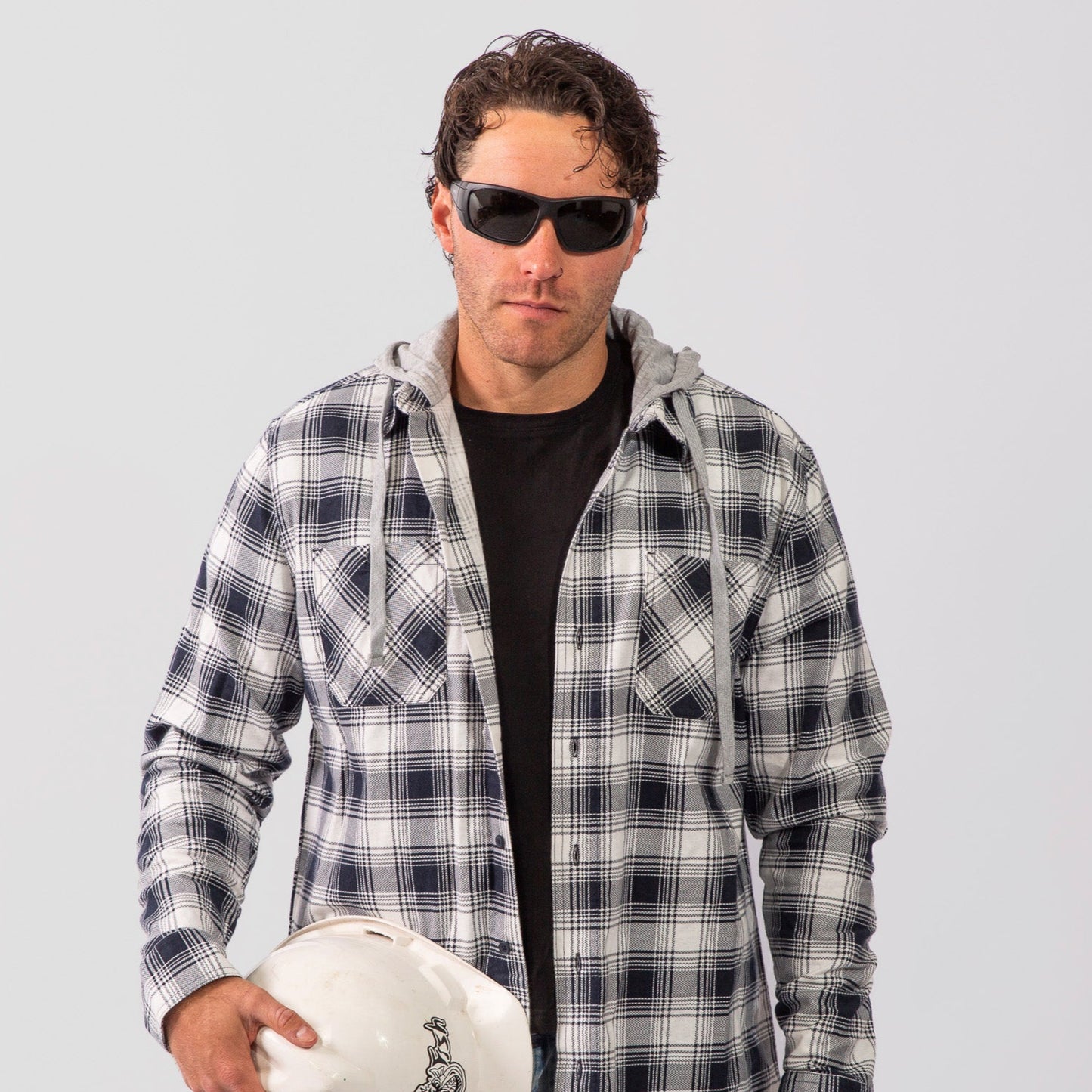 Man wearing a plaid shirt and Ugly Fish Safety sunglasses holding a hard hat against a white background
