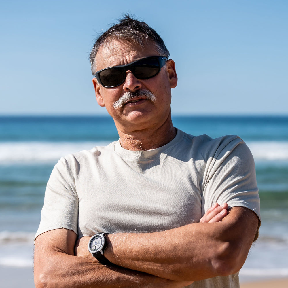 Man standing on a beach with arms crossed, wearing Ugly Fish polarised sunglasses and a white t-shirt.