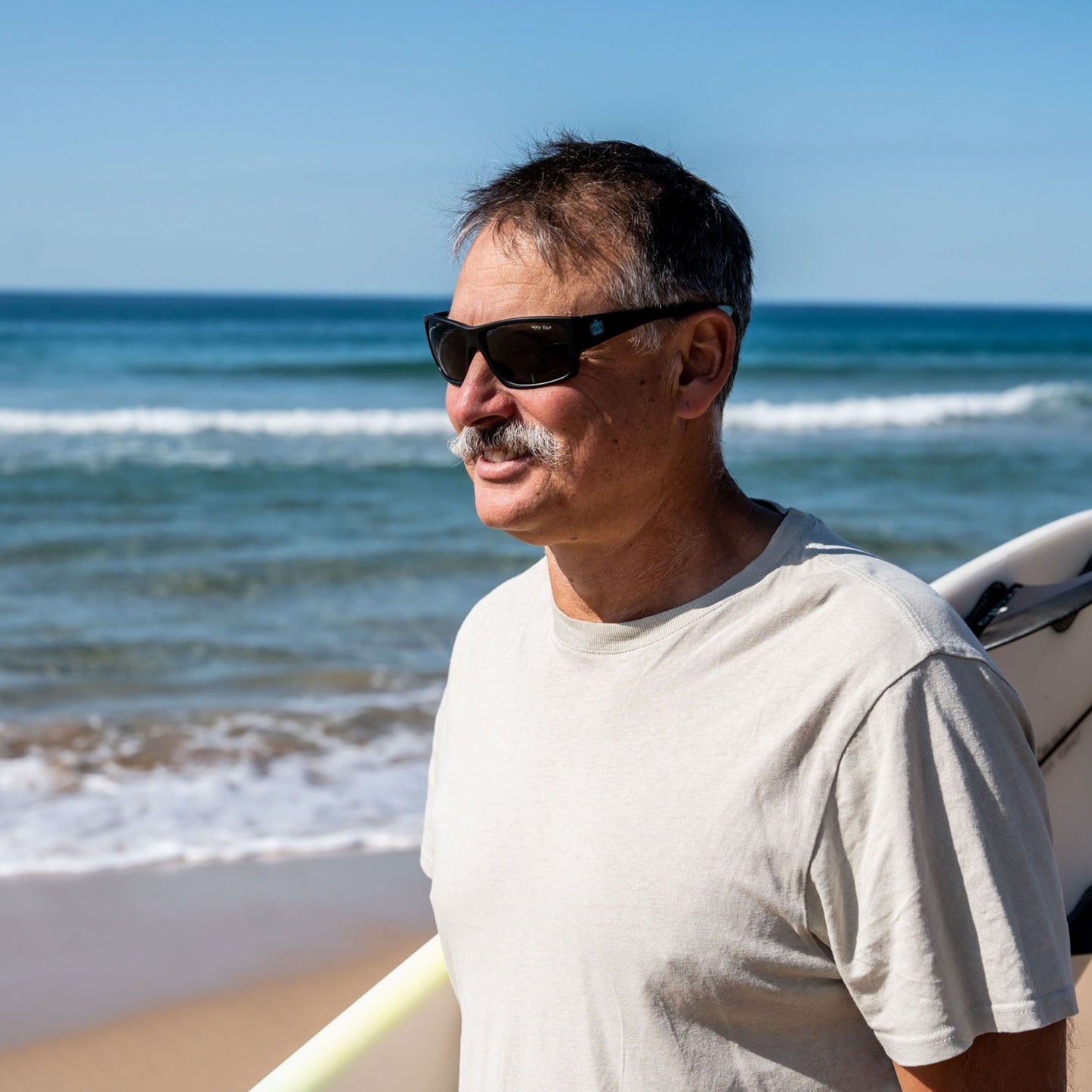 Man with surfboard wearing Ugly Fish floating sunglasses on a beach under a clear blue sky.