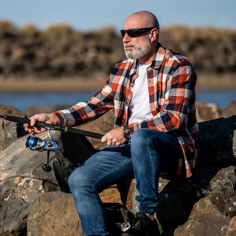 Man sitting on rocks by a body of water, wearing Ugly Fish polarised sunglasses holding a fishing rod.