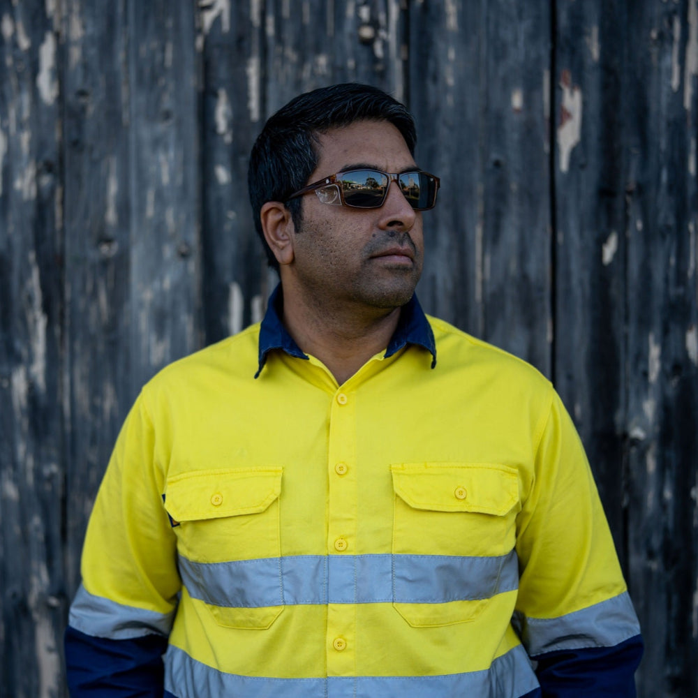 Man wearing a high-visibility yellow shirt and Ugly Fish Safety sunglasses with reflective stripes against a dark wooden background.