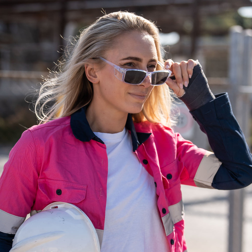 Woman wearing a pink jacket with black and white accents and Ugly Fish Safety sunglasses, holding a helmet.