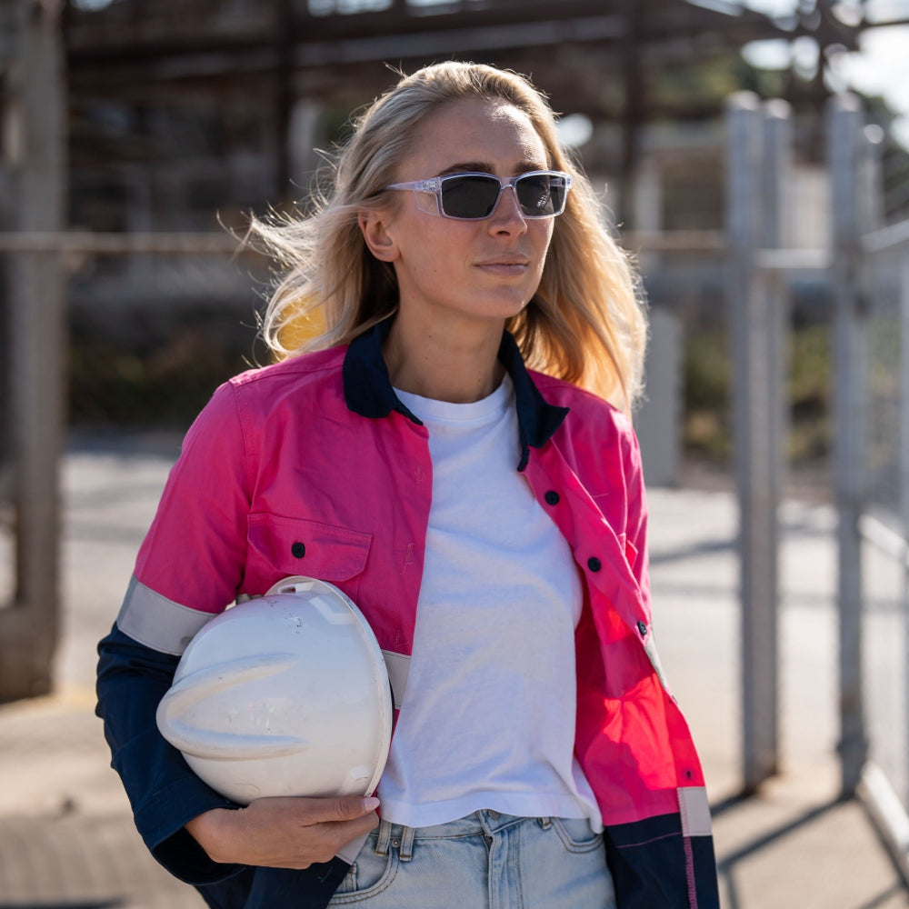 Woman wearing a pink jacket and Ugly Fish Safety sunglasses, holding a helmet outdoors.