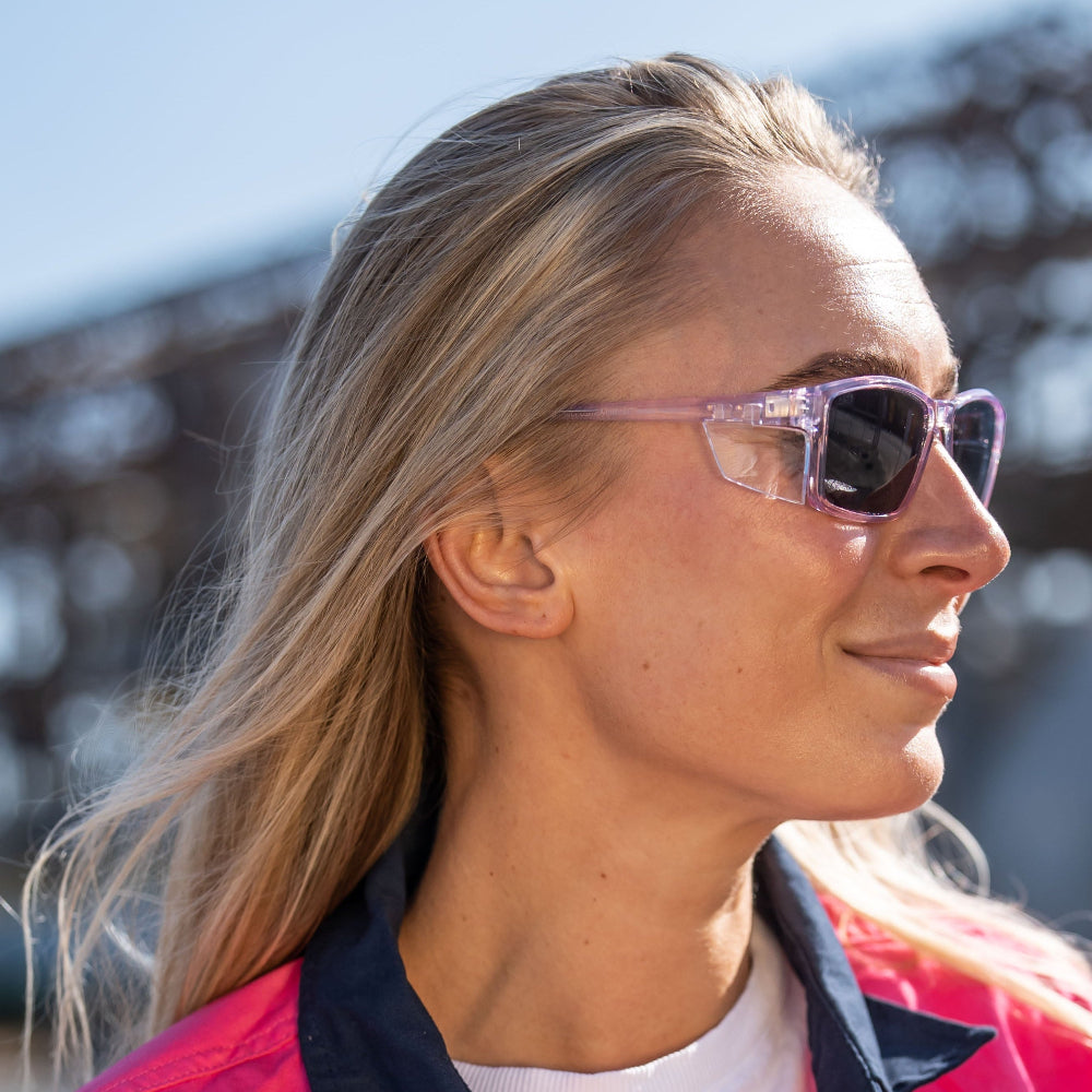 Woman wearing pink Ugly Fish Safety sunglasses and a red jacket with a blurred background.