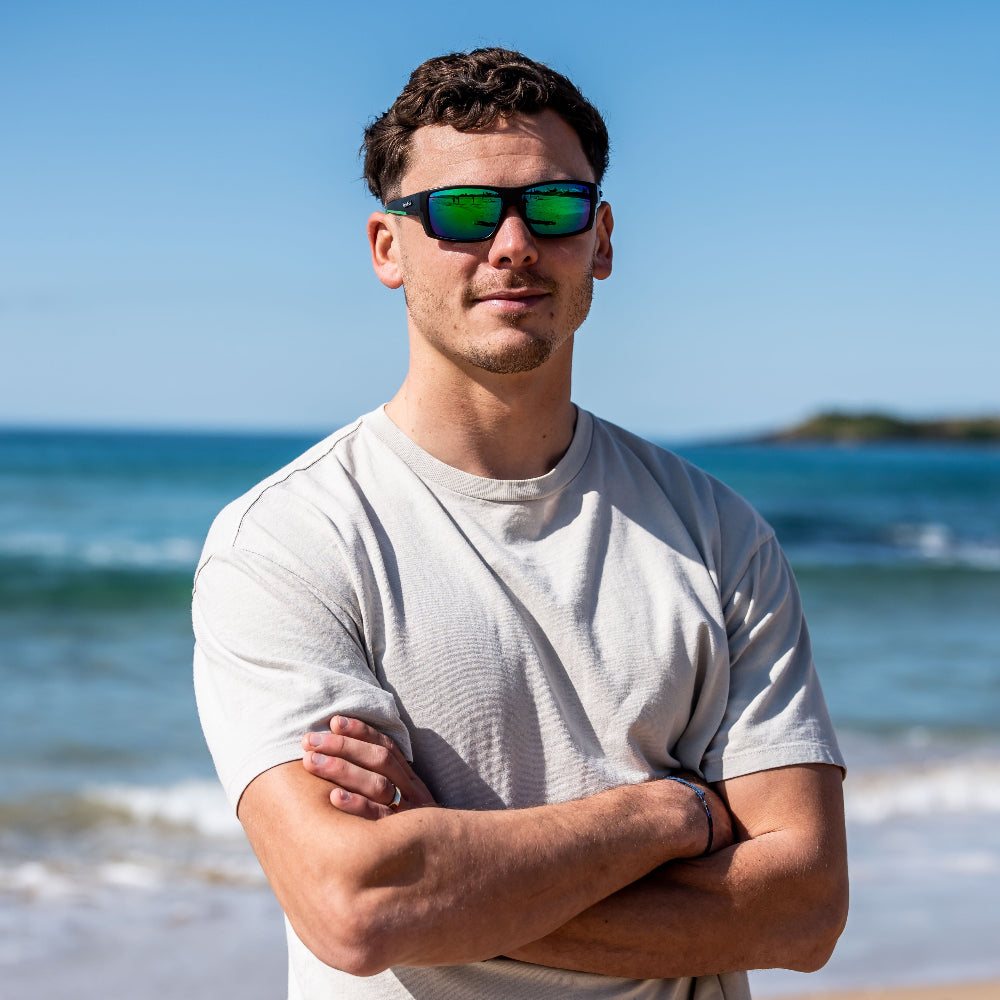 Man wearing Ugly Fish polarised sunglasses and a white t-shirt standing on a beach with ocean in the background