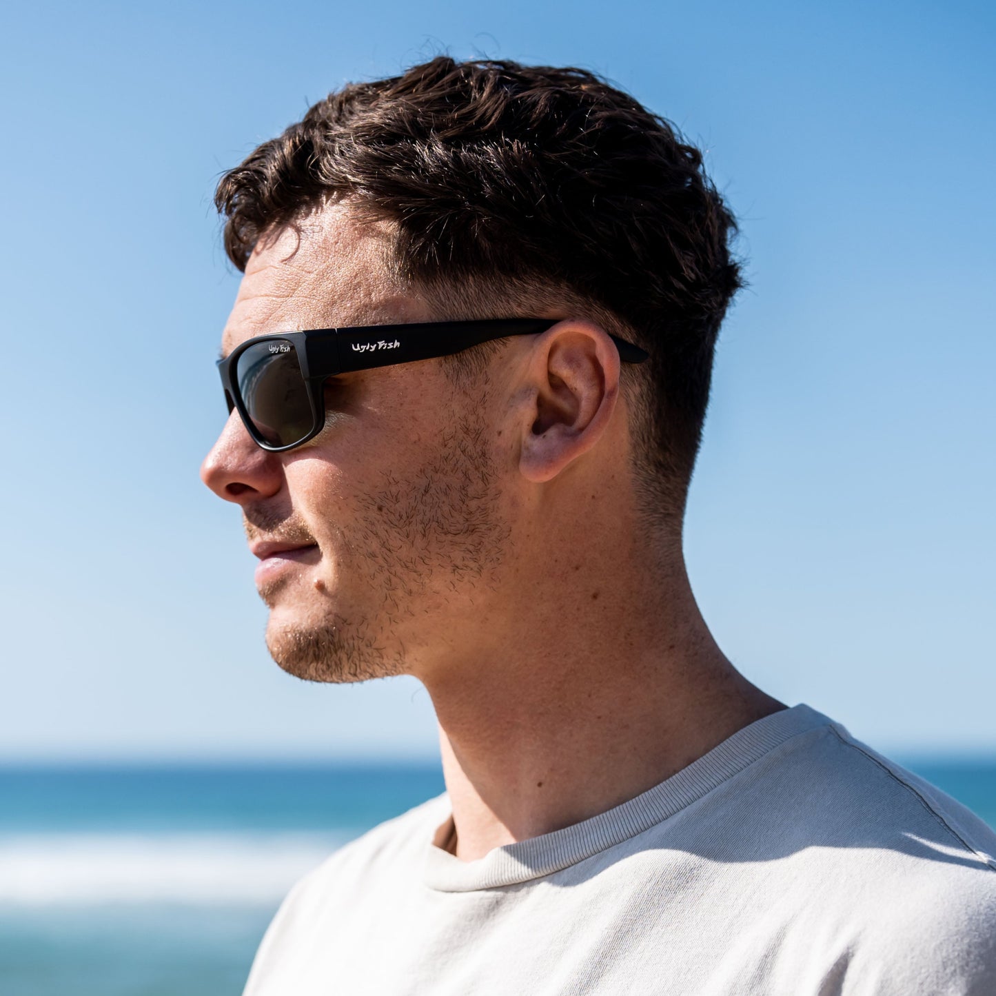 Man wearing Ugly Fish polarised sunglasses and a white t-shirt standing on a beach with clear blue sky and ocean.