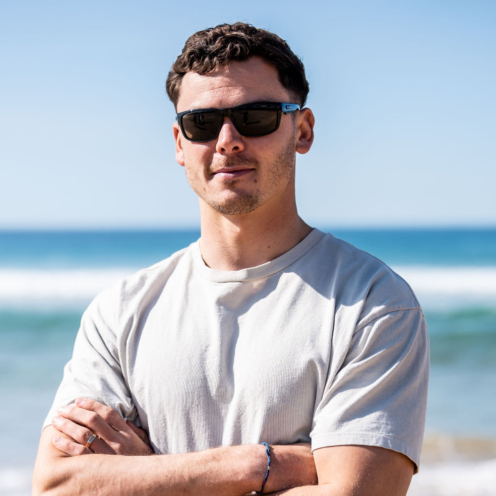 Man wearing Ugly Fish polarised sunglasses and a white t-shirt standing on a beach with ocean waves in the background