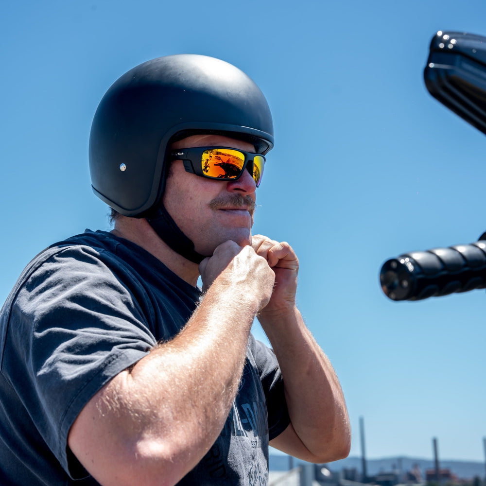 Man wearing a helmet and Ugly Fish motorcycle sunglasses by a waterfront with clear blue sky