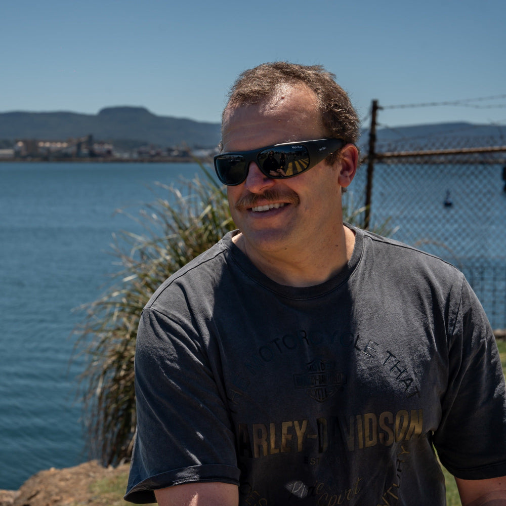 Man wearing Ugly Fish polarised sunglasses and a black t-shirt with text, standing by a body of water with mountains in the background.