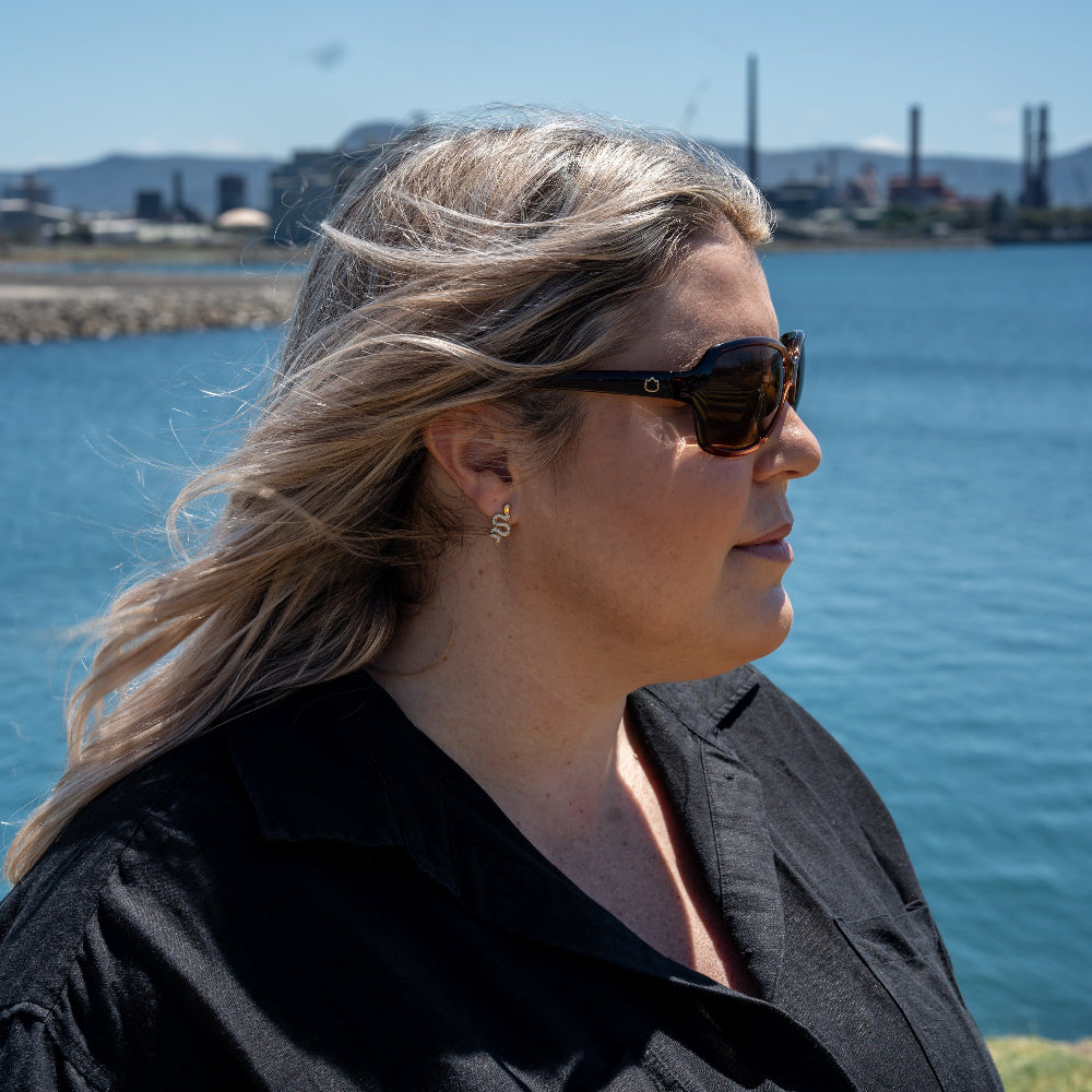 Woman wearing Ugly Fish polarised sunglasses and a black shirt by a body of water with industrial structures in the background.