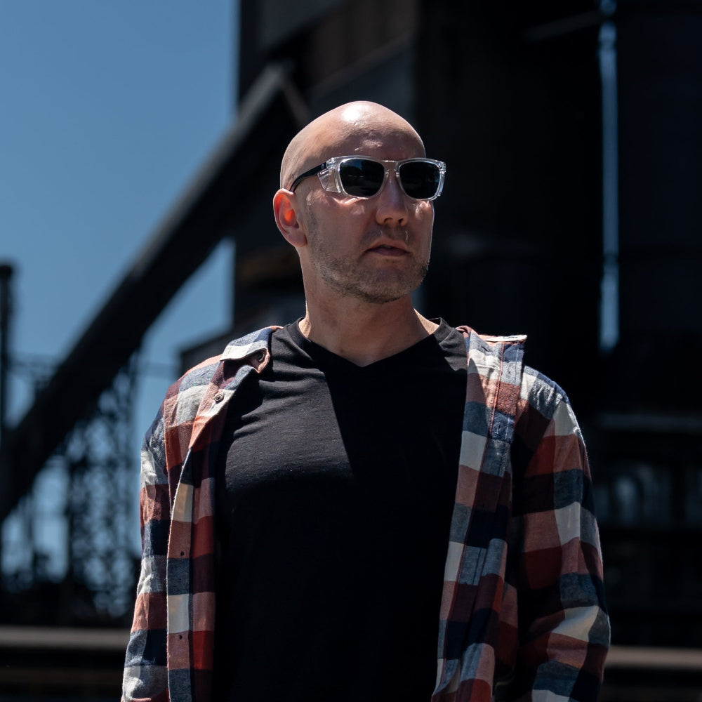 Man wearing Ugly Fish Safety sunglasses and a plaid shirt standing in front of industrial structures.