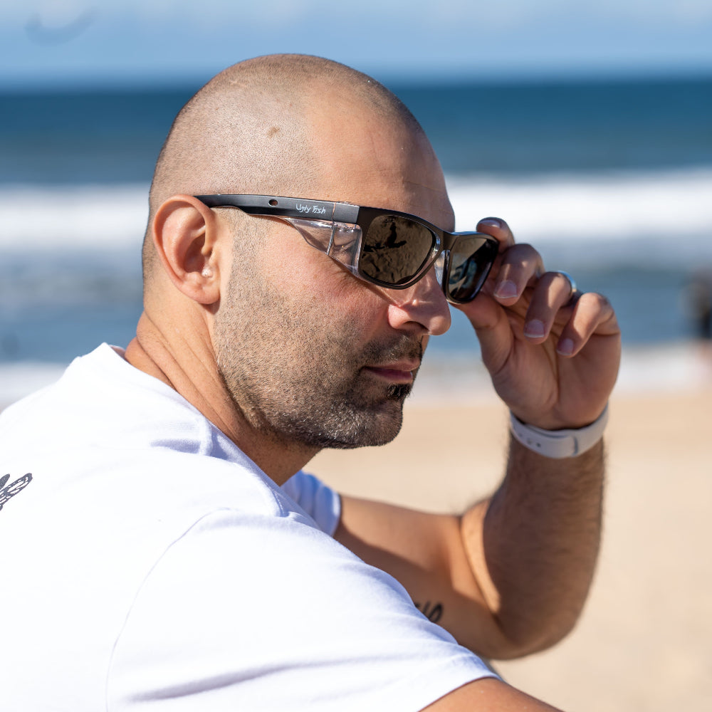 Man adjusting Ugly Fish Safety sunglasses on a beach with ocean in the background