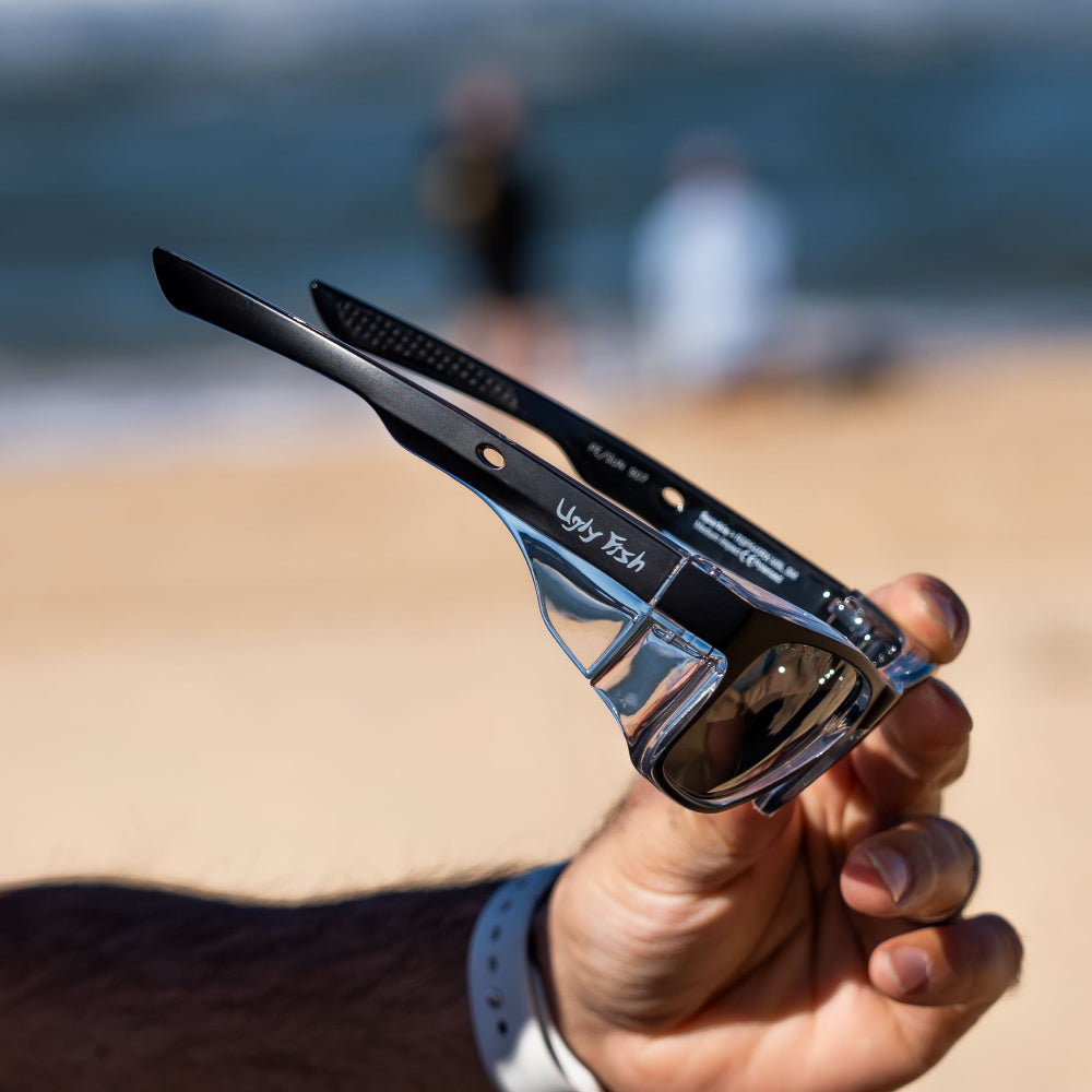 Person holding a pair of Ugly Fish Safety sunglasses with a blurred beach background.
