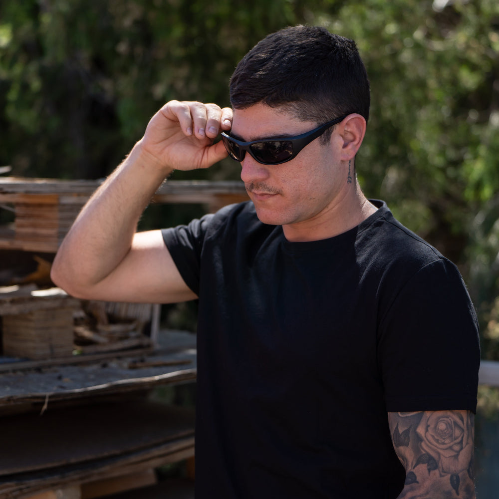 Man adjusting Ugly Fish Safety sunglasses outdoors with wooden pallets and greenery in the background.