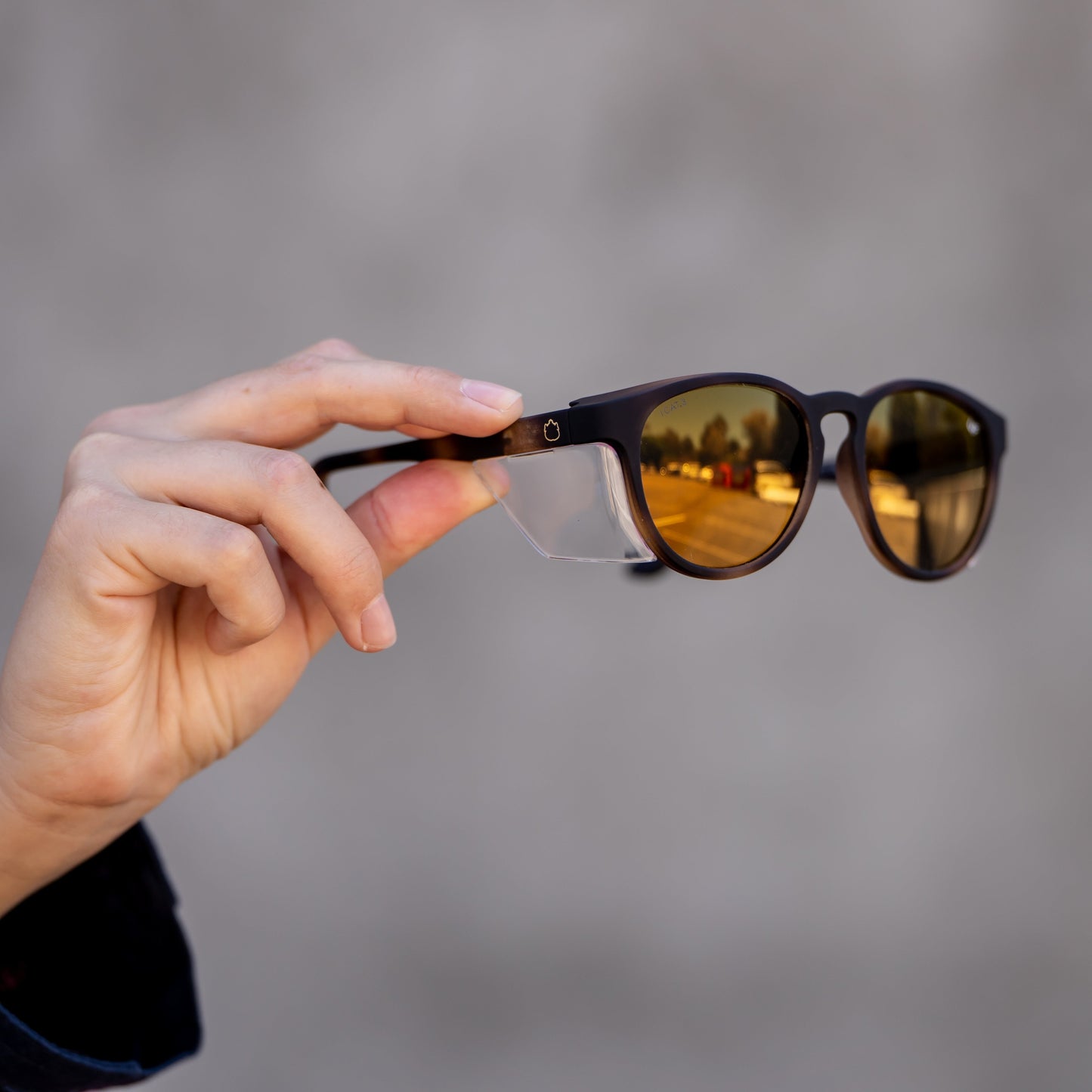 a hand holds a pair of brown ugly fish sunglasses with safety shields against a blurred gray background