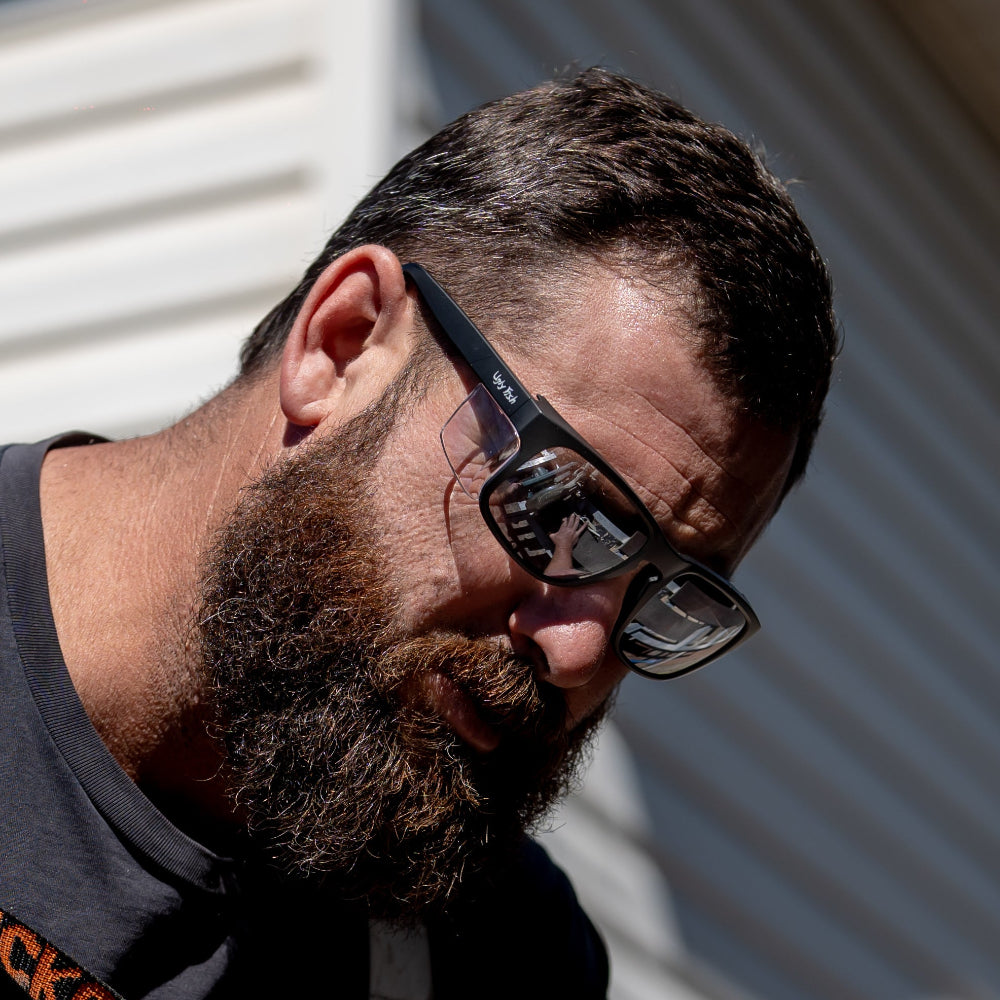 Man with a beard and Ugly Fish Safety glasses looking down, wearing a black shirt with visible text.