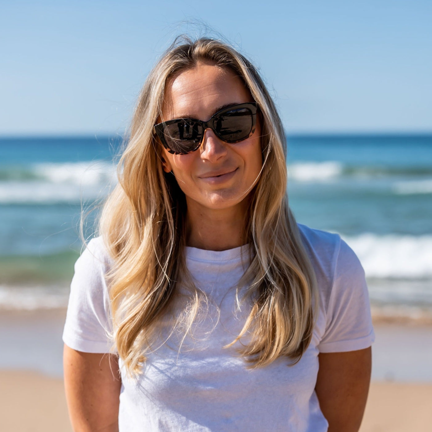 Woman standing on a beach wearing Ugly Fish polarised sunglasses and a white t-shirt with blue jeans.