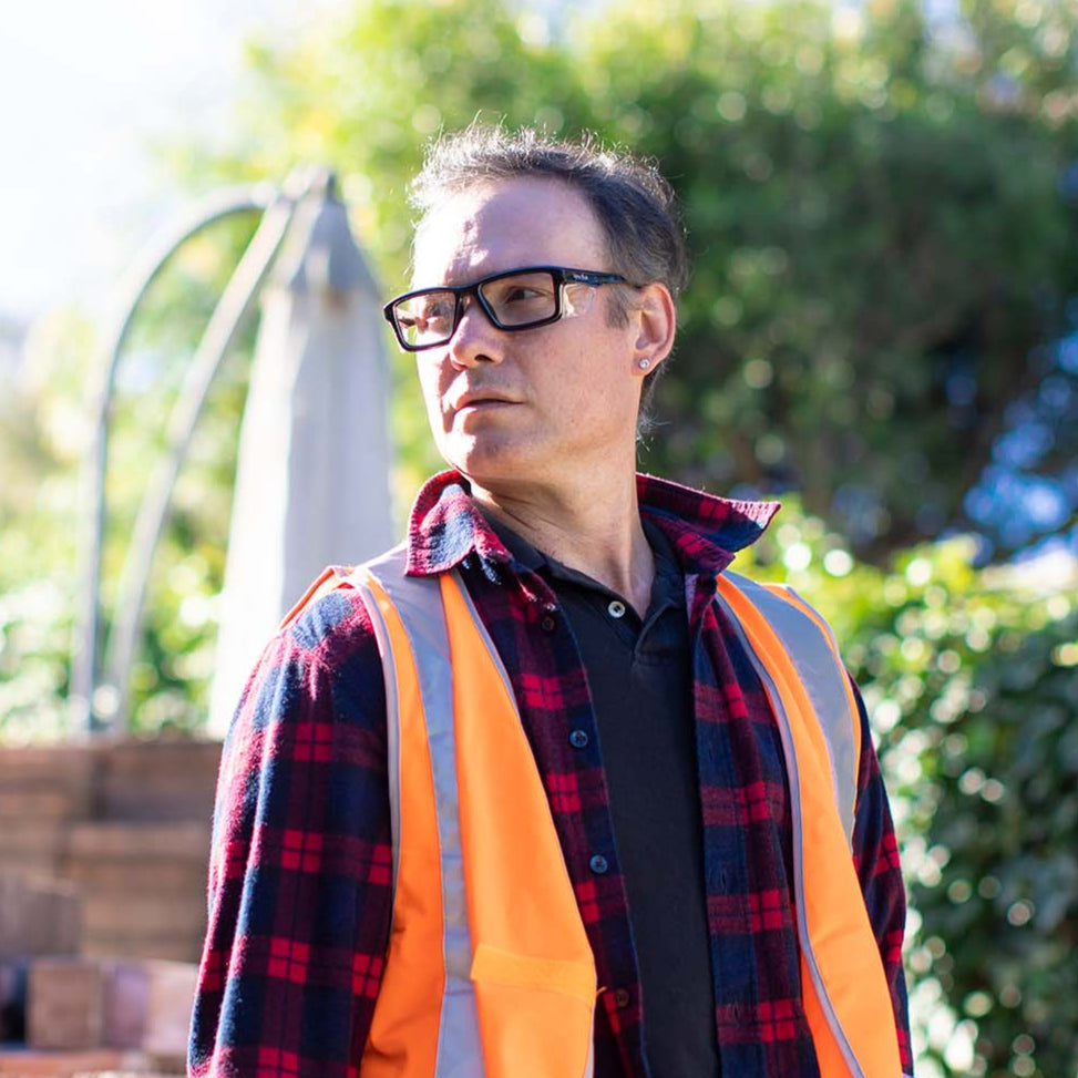Man wearing an orange safety vest and Ugly Fish safety glasses standing outdoors with greenery in the background