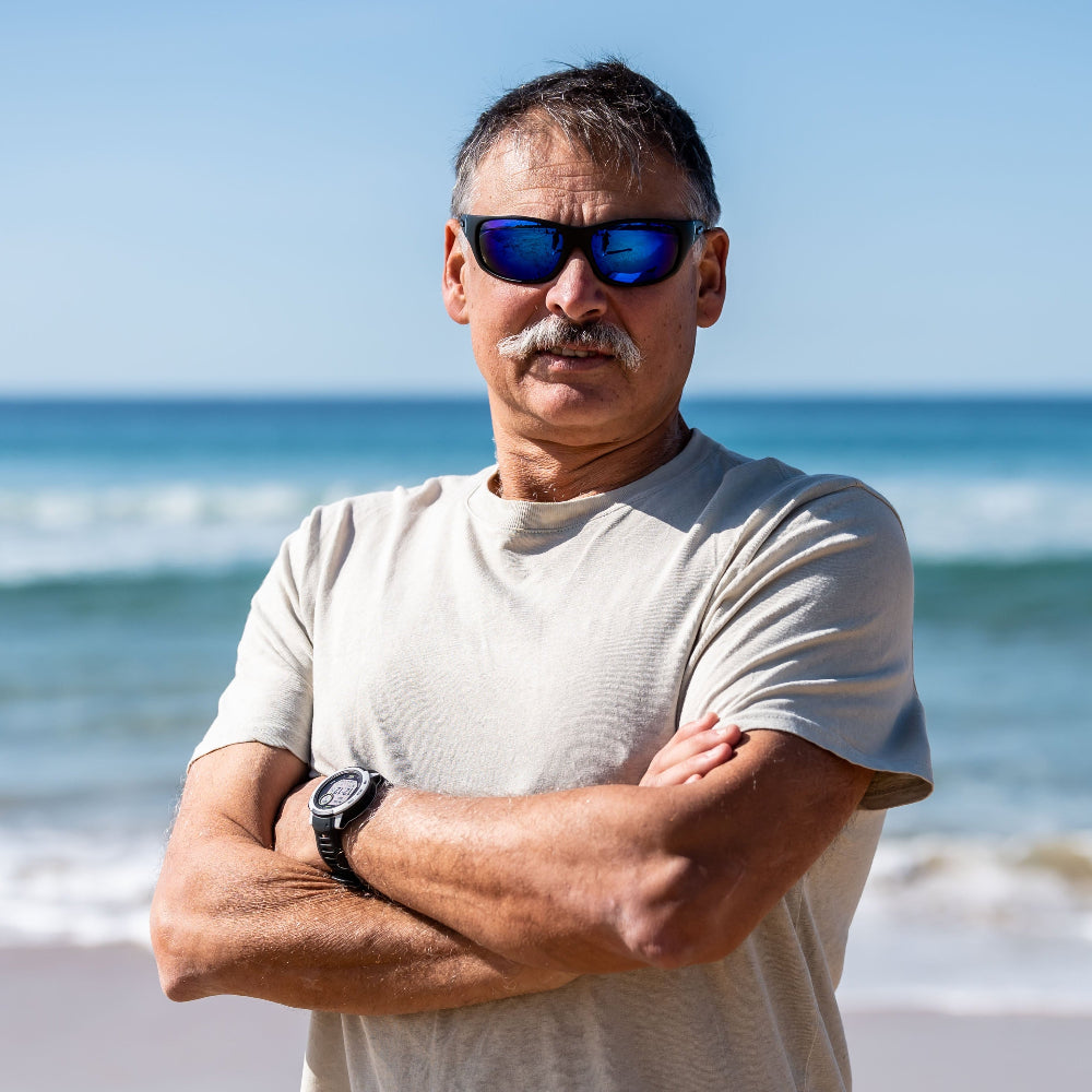 Man wearing sunglasses and a beige shirt standing on a beach with ocean waves in the background