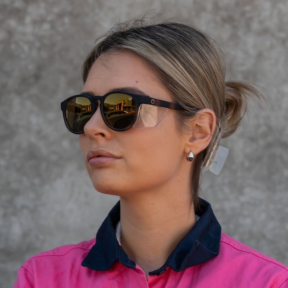 Woman wearing Ugly Fish Safety sunglasses and a pink shirt against a gray wall.