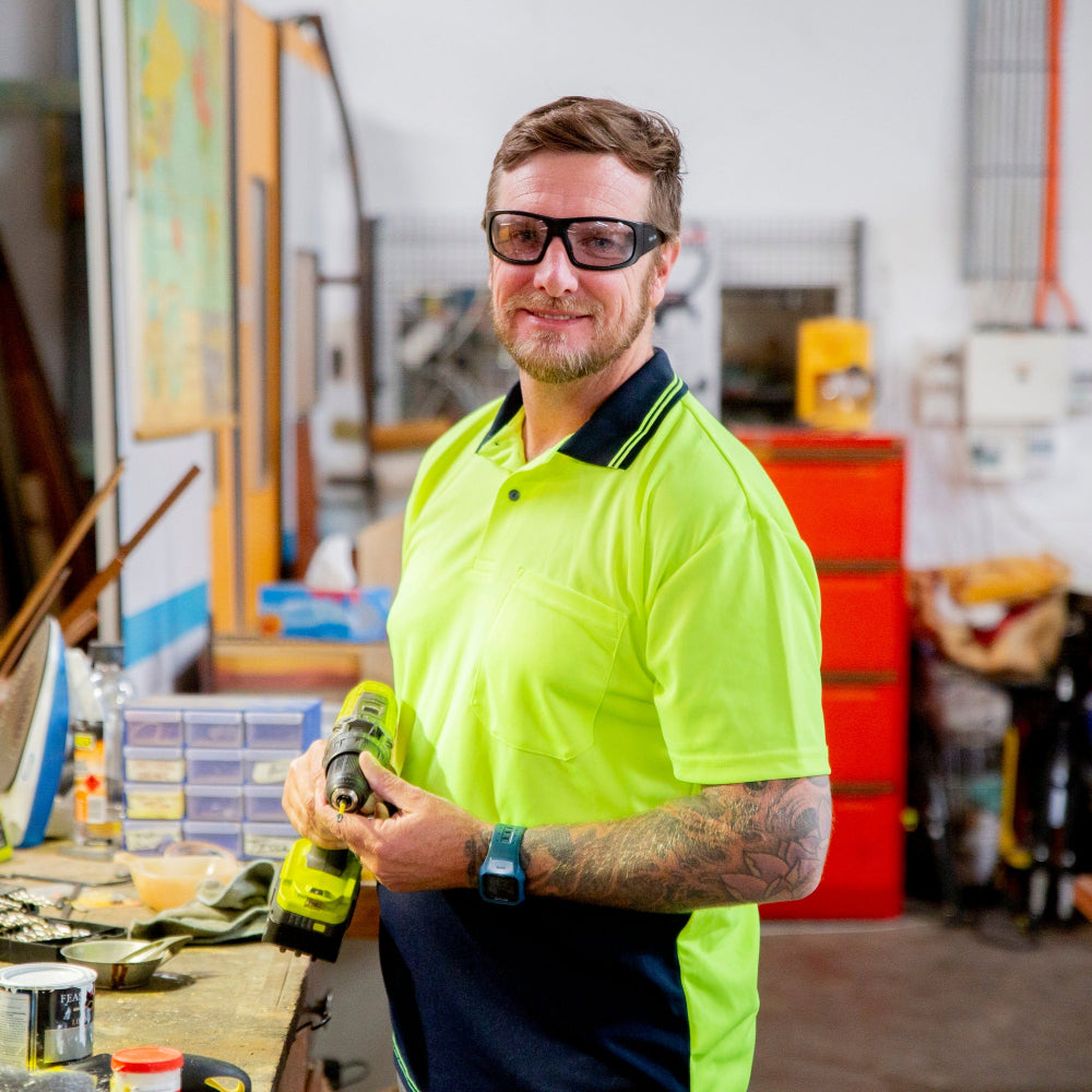 Man in a bright yellow safety shirt wearing Ugly Fish Safety sunglasses holding a drill in a workshop.