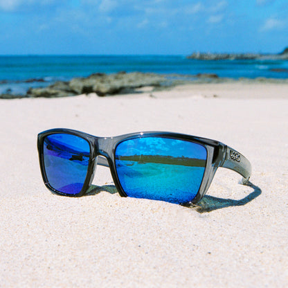 Ugly Fish polarised sunglasses with blue lenses on a sandy beach with ocean in the background