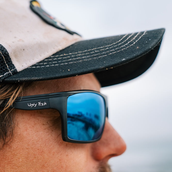 Person wearing Ugly Fish sunglasses with blur mirror lens and a cap against a plain background. 