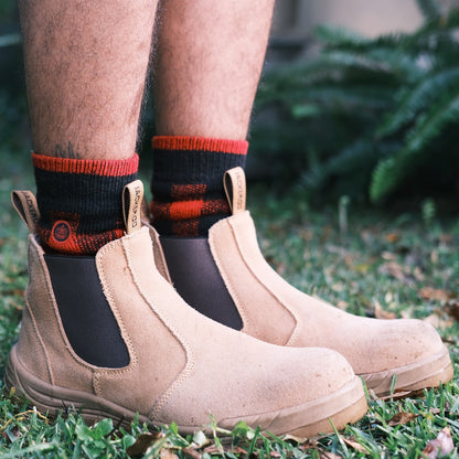 Person wearing beige work boots and black and red Ugly Fish socks on grass.