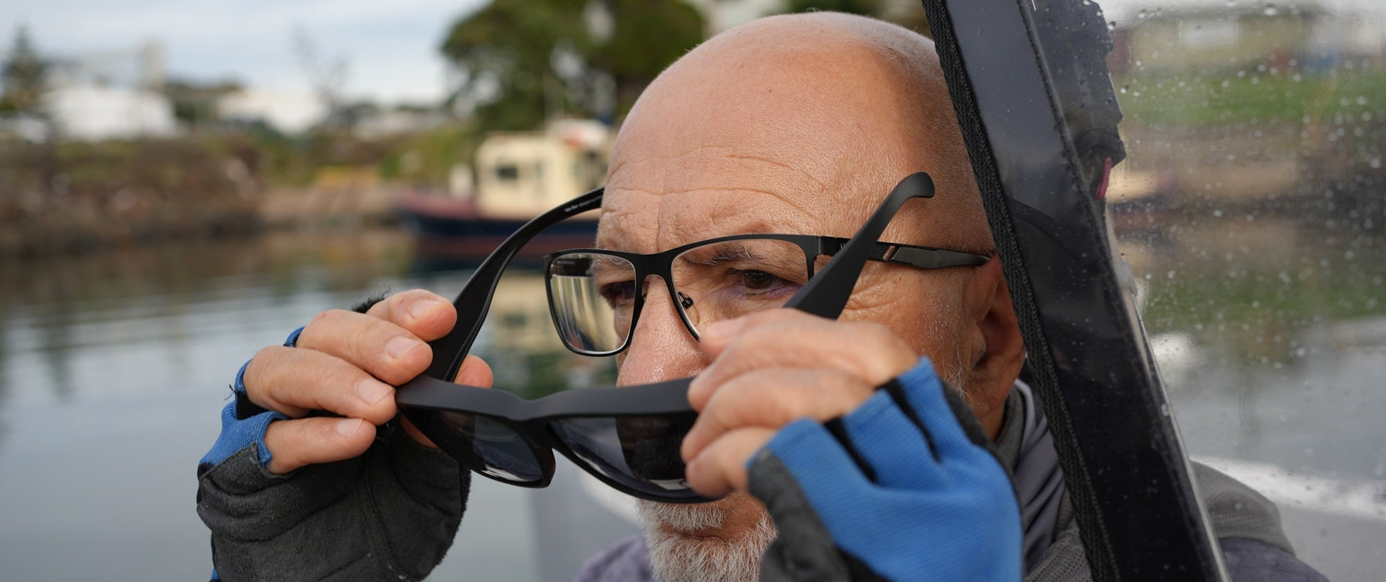 A close-up lifestyle shot of a bald man with a gray beard wearing black-framed specs and blue-and-black gloves. Putting a pair of Ugly Fish Fit Over sunglasses over the top while sitting by a body of water.