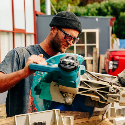 Man using a power tool outdoors with a building and trees in the background