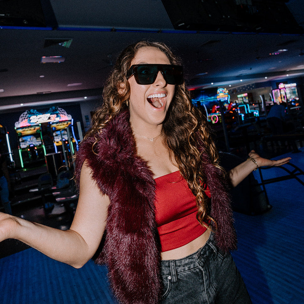Woman in a red top and maroon vest with Bohemian Retro sunglasses in an arcade setting