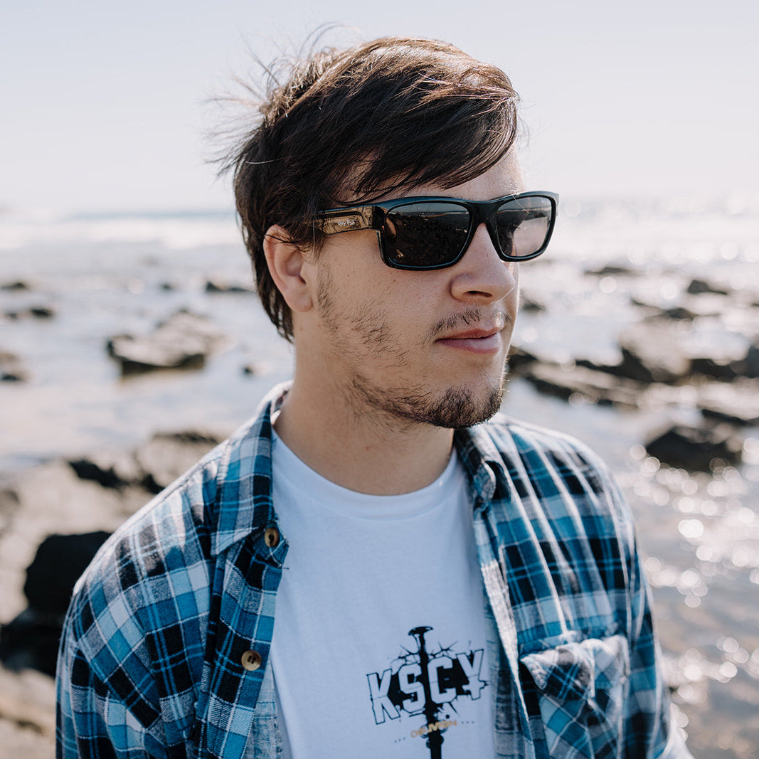 Man wearing Ugly Fish sunglasses and a plaid shirt standing by a body of water.