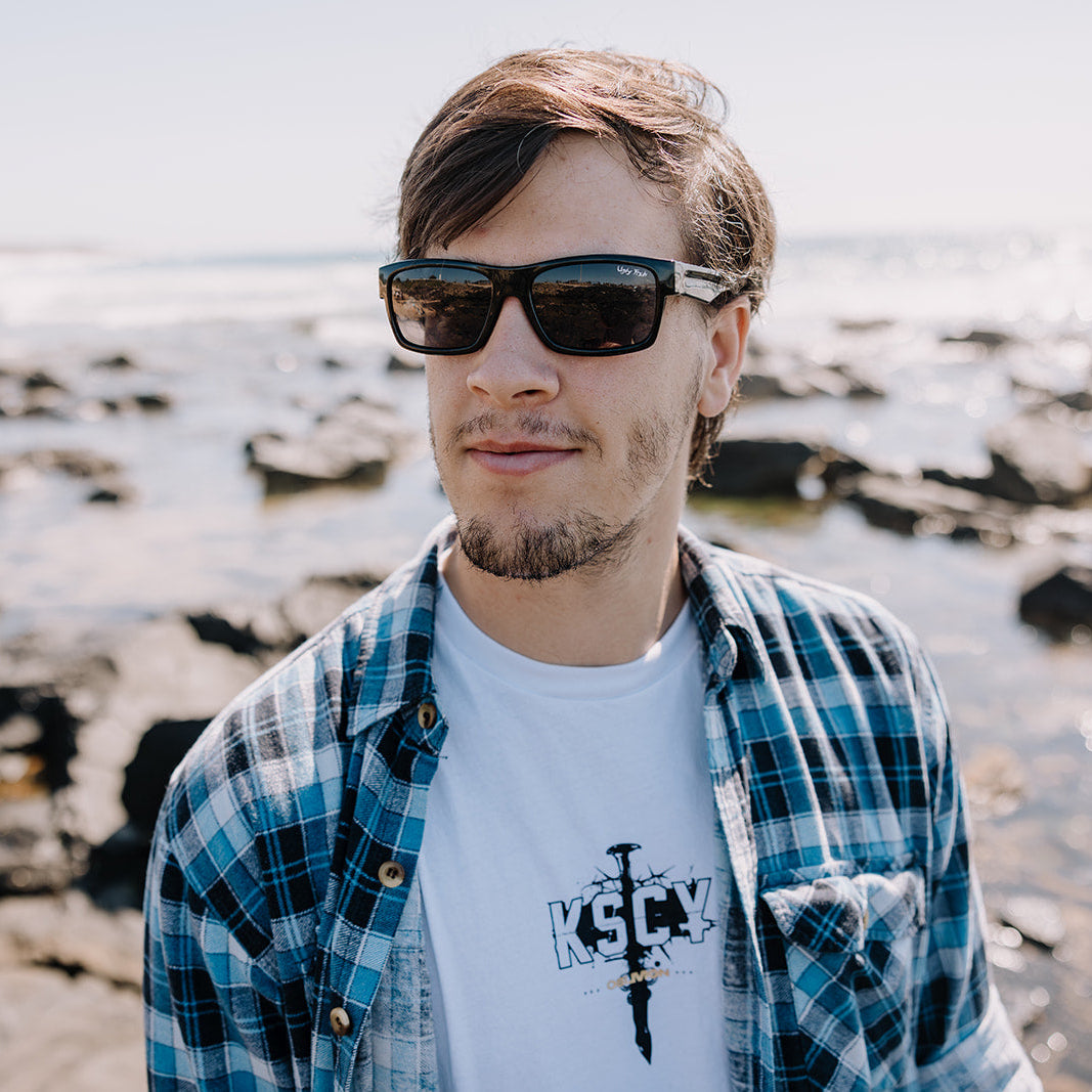 Man wearing Ugly Fish sunglasses and a plaid shirt with a white t-shirt underneath, standing on a rocky beach.