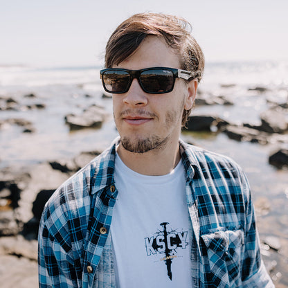 Man wearing Ugly Fish sunglasses and a plaid shirt with a white t-shirt underneath, standing on a rocky beach.