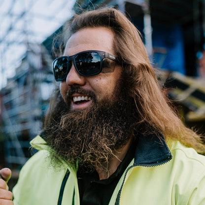 Man with long hair and beard wearing Ugly Fish Freestone safety sunglasses and a high-visibility jacket.
