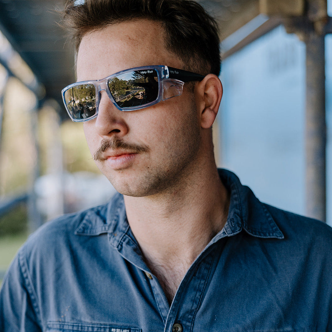 A man wearing Ugly Fish Cyclone Safety sunglasses with a blue denim shirt, photographed in a real-world setting with a blurred background.