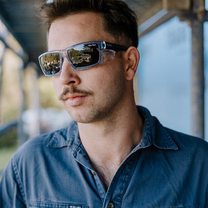 A man wearing Ugly Fish Cyclone Safety sunglasses with a blue denim shirt, photographed in a real-world setting with a blurred background.