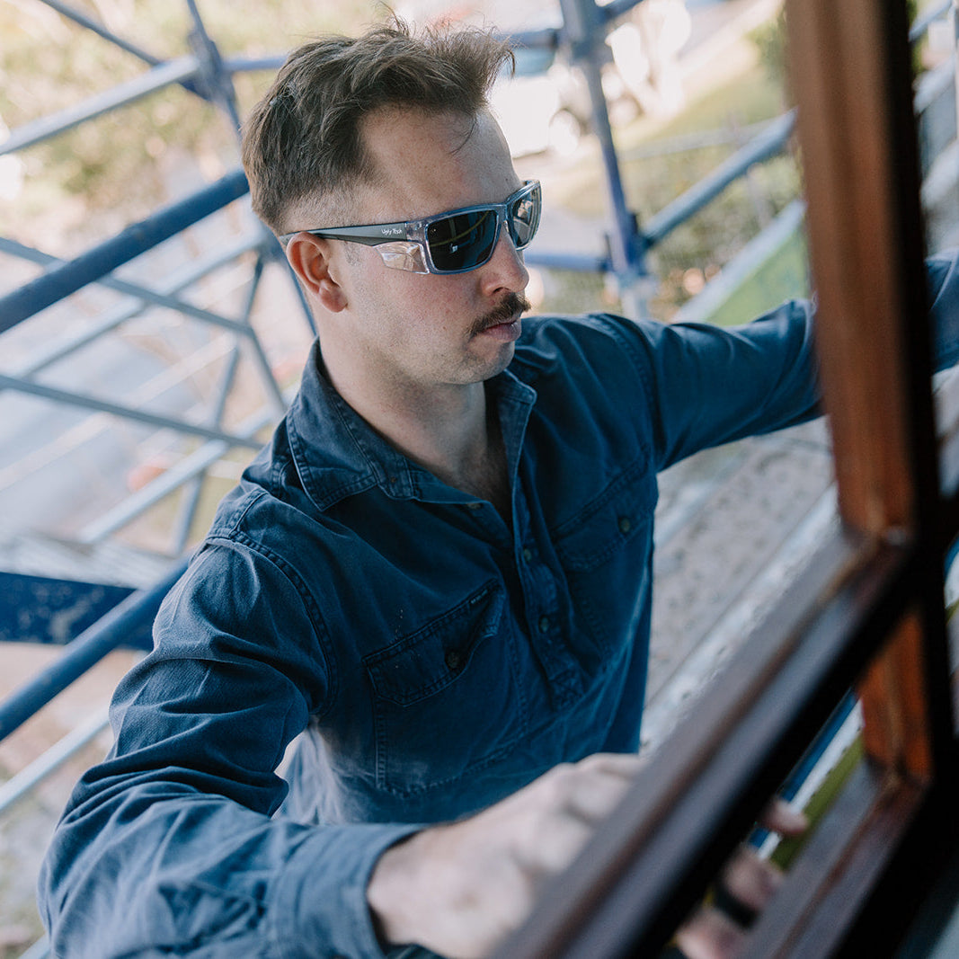 A man wearing a dark blue button-up shirt and Ugly Fish Cyclone safety sunglasses looks out from behind a wooden window frame, with a metal structure and blurred outdoor background.