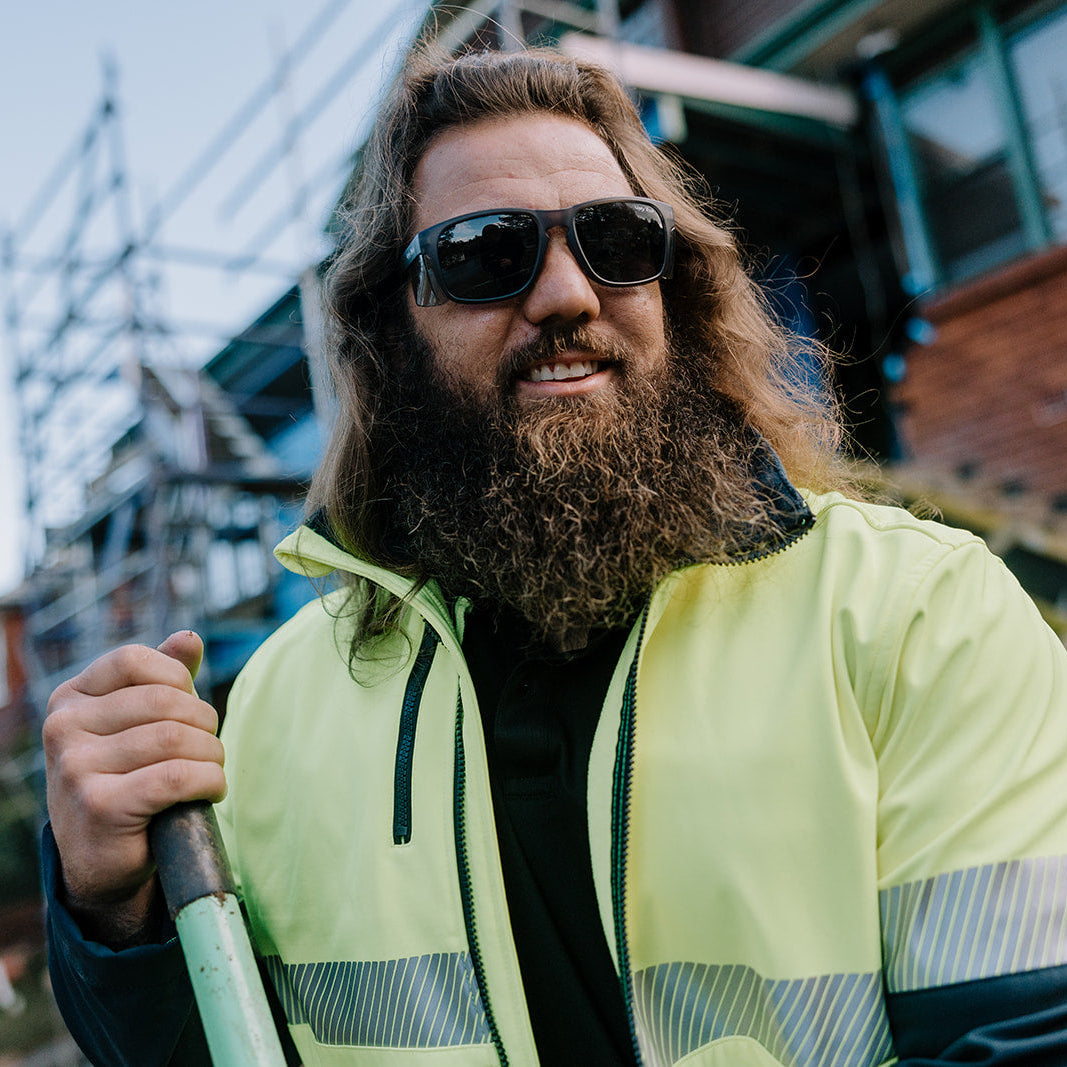 Man with long hair and beard wearing Ugly Fish Freestone Safety sunglasses and a high-visibility jacket outdoors.