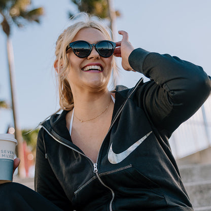 A woman sitting on outdoor concrete steps wearing Ugly fish women's sunglasses, a black zip-up hoodie and matching sweatpants, holding a coffee cup under her arm. Palm trees and a blue sky are visible in the background.